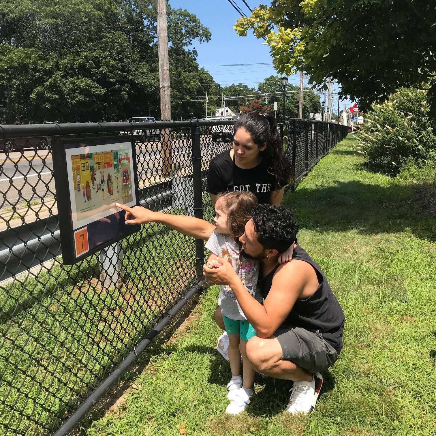 A family reading a StoryWalk panel mounted to a chain-link fence